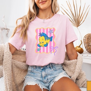 A woman wearing a pink t-shirt with a cartoon fish graphic and denim shorts is sitting in a chair.