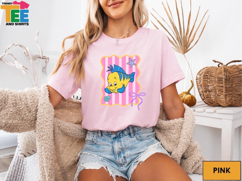 A woman wearing a pink t-shirt with a cartoon fish graphic and denim shorts is sitting in a chair.
