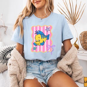A woman wearing a light blue t-shirt with a cartoon fish graphic and denim shorts is sitting on a chair.
