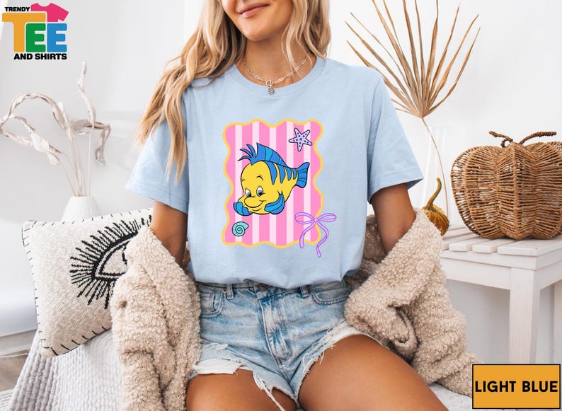A woman wearing a light blue t-shirt with a cartoon fish graphic and denim shorts is sitting on a chair.