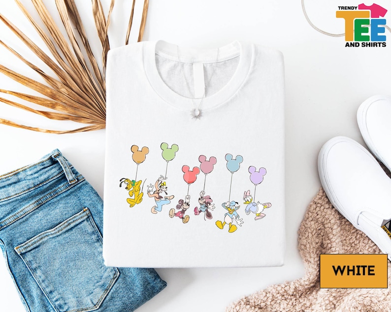 a white t-shirt with a colorful graphic of Disney characters, including Mickey Mouse, Donald Duck, Goofy, Minnie Mouse, and Donald Duck, along with balloons. The t-shirt is laid out on a white surface, along with a pair of blue jeans, white sneakers, and a beige blanket.