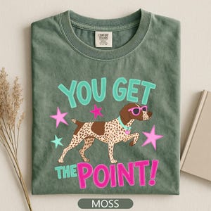 May include: A moss green t-shirt featuring a graphic of a brown and white spotted dog wearing bright pink sunglasses and a light blue collar. The text reads YOU GET THE POINT! in light blue and pink, surrounded by pink and light blue stars.