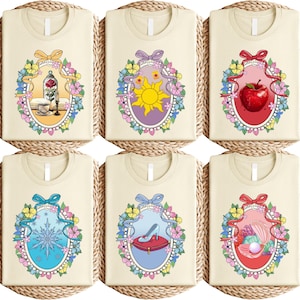 May include: Six cream-colored t-shirts with different fairytale-themed designs. Each shirt features a colorful oval frame with floral and ribbon accents. The designs include a rose, sun, apple, snowflake, glass slipper, and seashell.