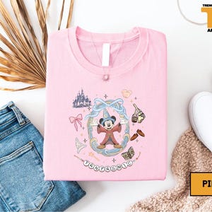 May include: Pink crew neck t-shirt featuring a cartoon Mickey Mouse design with the word "Fantastic" below. The design includes a castle, bow, and other magical elements. The shirt is displayed with blue jeans and white shoes.