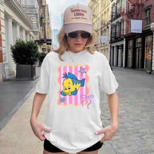 A woman wearing a white t-shirt with a cartoon fish graphic and a baseball cap stands on a city street.