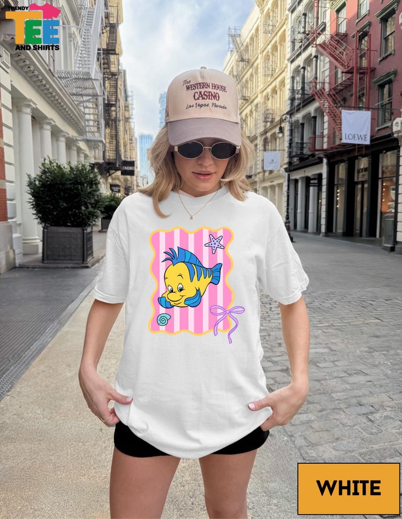 A woman wearing a white t-shirt with a cartoon fish graphic and a baseball cap stands on a city street.