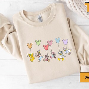 A beige sweatshirt with a graphic design featuring a group of cartoon characters, including Mickey Mouse, Donald Duck, Goofy, and Minnie Mouse, along with balloons.