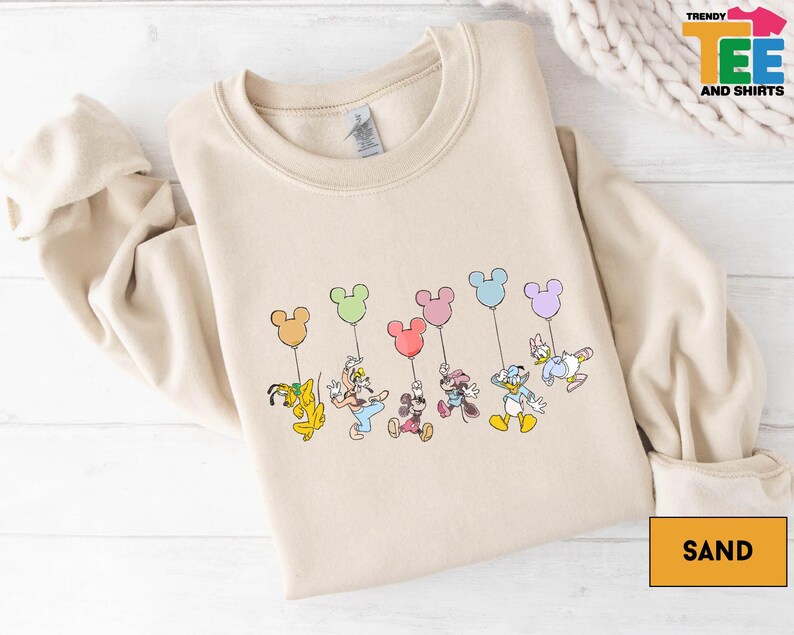 A beige sweatshirt with a graphic design featuring a group of cartoon characters, including Mickey Mouse, Donald Duck, Goofy, and Minnie Mouse, along with balloons.