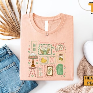 May include: Heather peach-colored t-shirt with a graphic design featuring a hen in a basket, a duck, a lamp, and other vintage-style illustrations. The text "Granny Era" is visible. The shirt is paired with blue jeans and white sneakers.