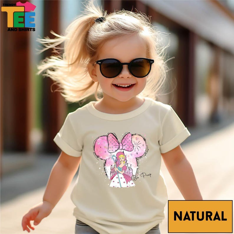 A young girl wearing a white t-shirt with a cartoon character and sunglasses, smiling and walking outdoors.
