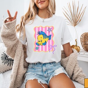 A woman wearing a white t-shirt with a cartoon fish graphic and a peace sign, sitting on a couch with a beige jacket draped over her.