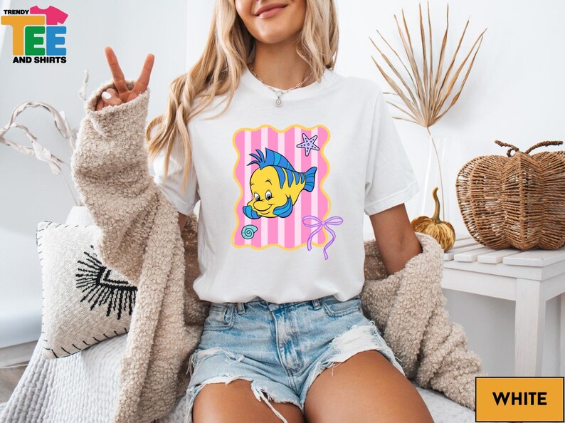 A woman wearing a white t-shirt with a cartoon fish graphic and a peace sign, sitting on a couch with a beige jacket draped over her.