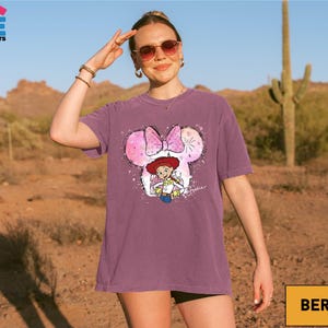 A woman wearing sunglasses and a purple t-shirt with a cartoon character is standing in a desert-like environment.