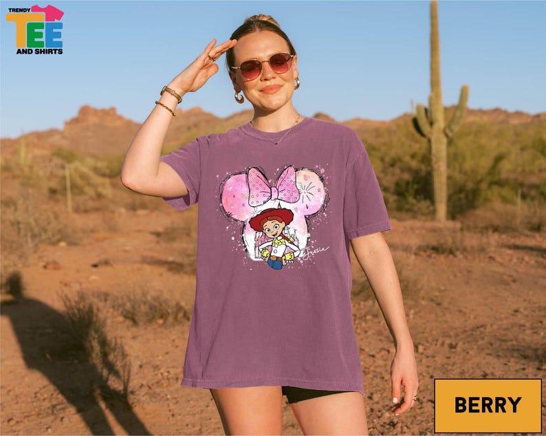 A woman wearing sunglasses and a purple t-shirt with a cartoon character is standing in a desert-like environment.