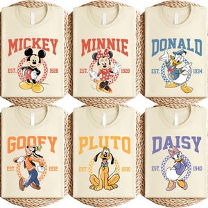 May include: Six cream-colored t-shirts featuring Disney characters. Each shirt displays a character's name in bold letters, along with the character's image and the year of establishment. Characters include Mickey, Minnie, Donald, Goofy, Pluto, and Daisy.