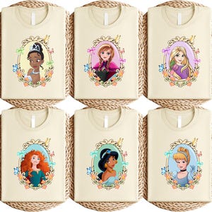 May include: Six cream-colored t-shirts featuring different princess illustrations within ornate frames. Each princess is depicted with vibrant colors and surrounded by floral and butterfly accents. The shirts are arranged on round, woven placemats.