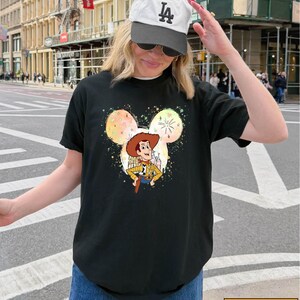 A woman wearing a black t-shirt with a cartoon character and a baseball cap is standing on a city street.