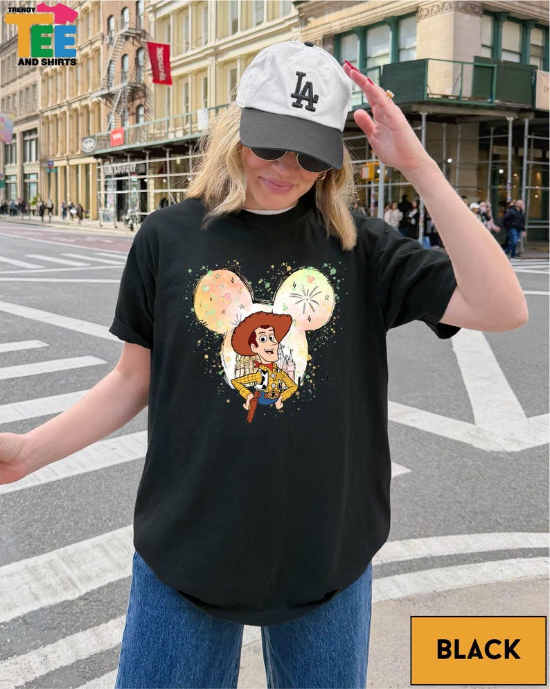 A woman wearing a black t-shirt with a cartoon character and a baseball cap is standing on a city street.