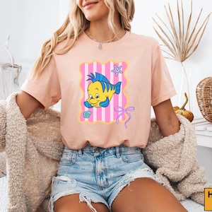 A woman wearing a pink t-shirt with a cartoon fish design and denim shorts is sitting in a room with a woven basket and a plant in the background.
