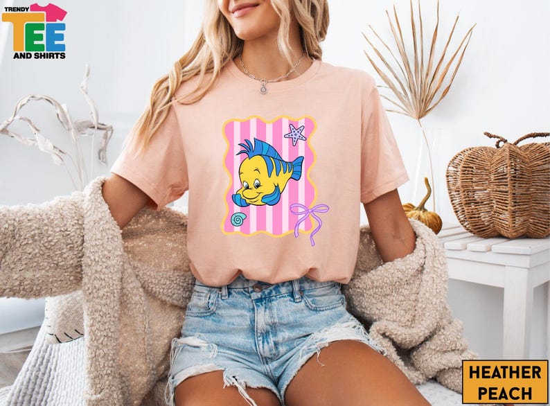 A woman wearing a pink t-shirt with a cartoon fish design and denim shorts is sitting in a room with a woven basket and a plant in the background.