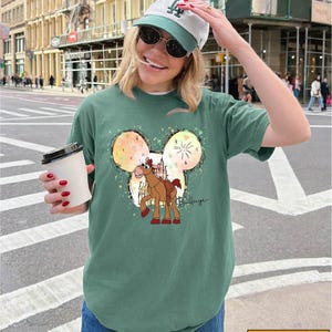 A woman wearing a green t-shirt with a Mickey Mouse design and a baseball cap is standing on a city street, holding a coffee cup.