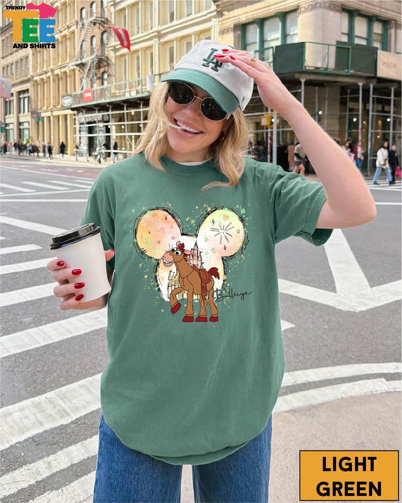 A woman wearing a green t-shirt with a Mickey Mouse design and a baseball cap is standing on a city street, holding a coffee cup.