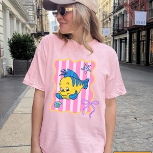 A woman wearing a pink t-shirt with a cartoon fish graphic and a pink ribbon, standing on a city street.