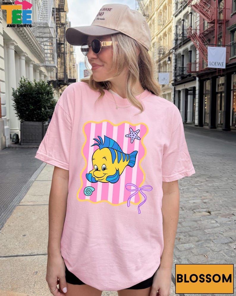 A woman wearing a pink t-shirt with a cartoon fish graphic and a pink ribbon, standing on a city street.