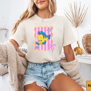 A woman wearing a white t-shirt with a cartoon fish graphic and denim shorts is sitting in a room with a woven basket and a plant.