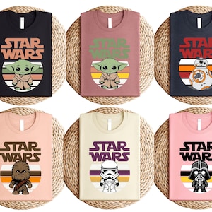 May include: Six t-shirts with Star Wars character graphics. The shirts are black, mauve, navy, peach, cream, and pink. Each shirt features a different character: Baby Yoda, BB-8, Chewbacca, a Stormtrooper, and Darth Vader, with the Star Wars logo.