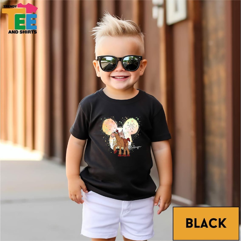 A young boy wearing a black t-shirt with a graphic of a horse and fireworks, smiling and standing in front of a wooden wall.