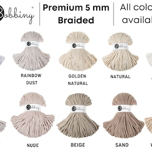 Może przedstawiać: R&oacute;żne rodzaje 5 mm plecionej sznurka bawełnianego w r&oacute;żnych kolorach, w tym biały, tęczowy pył, złoty naturalny, naturalny, kremowy, księżycowy, nude, beżowy, piaskowy i ciepły beż. Sznurek jest produkowany przez Bobbiny.