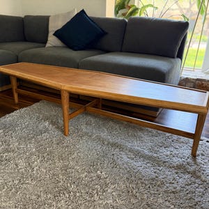 May include: A mid-century modern wooden coffee table with a rectangular top and angled legs. The table has a light brown finish and a lower shelf for storage. The table is approximately 60 inches long.
