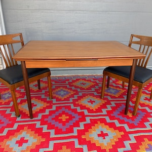 May include: A rectangular wooden dining table with a smooth top and four straight legs, accompanied by two wooden chairs featuring open backrests and black upholstered seats. The furniture set is arranged on a vibrant, patterned floor covering.