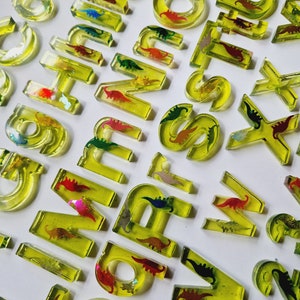 Peut inclure: Un ensemble de lettres de l'alphabet en tuiles jaunes translucides avec des silhouettes de dinosaures colorées intégrées. Les lettres sont en majuscules et sont disposées dans un ordre aléatoire.