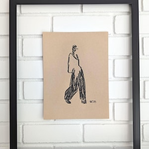 May include: A framed sketch of a figure in flowing trousers and a jacket, drawn in black on a tan background. The artwork is in a black frame, mounted on a white brick wall. The artist's signature is visible.