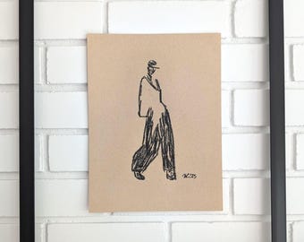 May include: A framed sketch of a person in loose-fitting clothing, drawn in black on tan paper. The artwork is in a black frame, mounted on a white brick wall. The artist's signature is in the bottom right corner.
