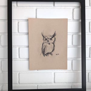 May include: A sketch of an owl in black ink on tan paper, framed in a black square frame. The owl has large eyes and detailed feathering. The artwork is displayed against a white brick wall.