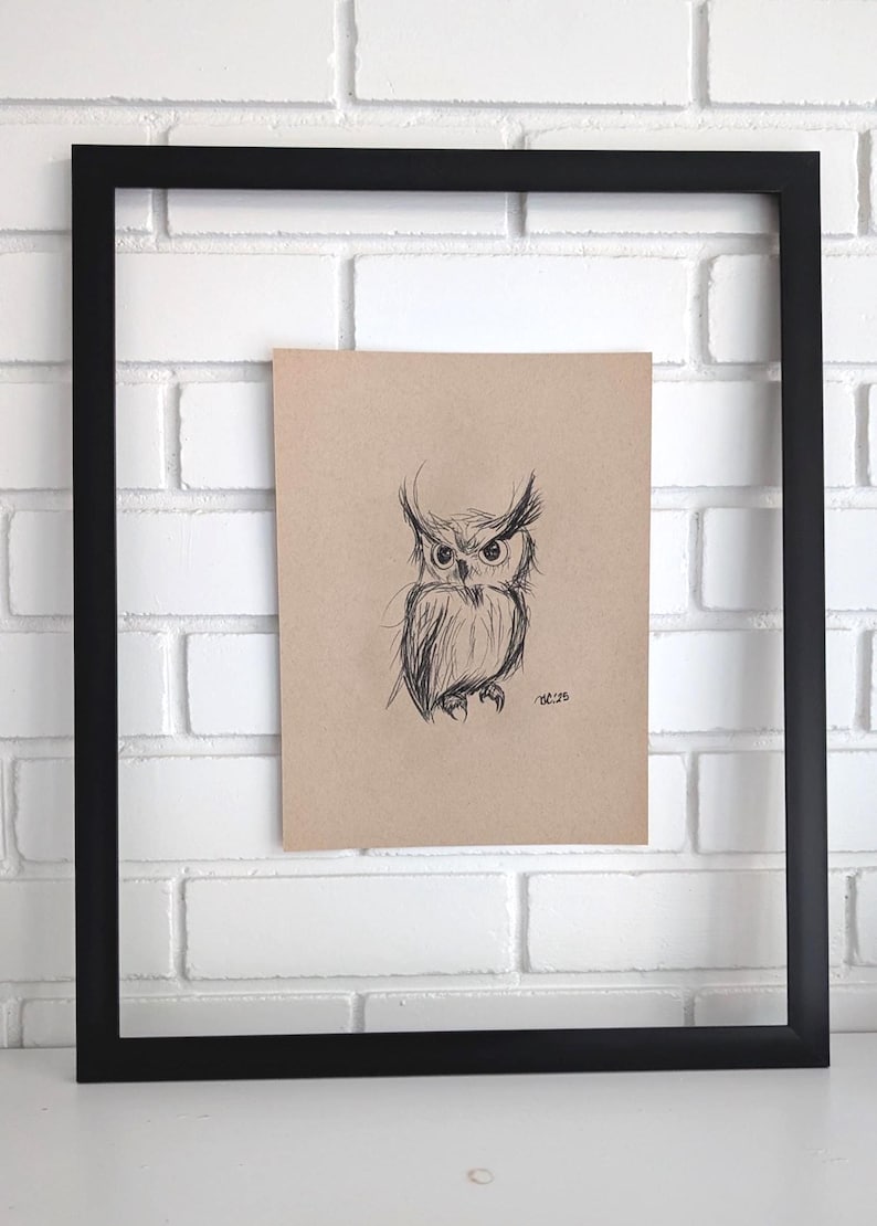 May include: A sketch of an owl in black ink on tan paper, framed in a black square frame. The owl has large eyes and detailed feathering. The artwork is displayed against a white brick wall.