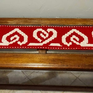 May include: A red and white crocheted table runner with a repeating pattern of white hearts and swirls. The runner is on a wooden table with a glass top.