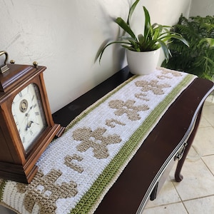 Könnte beinhalten: Ein gehäkelter Tischläufer mit weißem Hintergrund und einem Muster aus braunen Lebkuchenmännern. Der Läufer hat grüne Ränder und wird auf einem dunkelbraunen Tisch präsentiert.