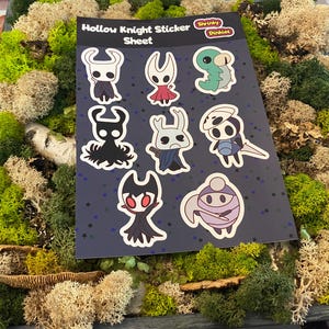 Peut inclure: Une planche d'autocollants Hollow Knight avec divers personnages du jeu. Les autocollants sont sur un fond bleu foncé avec des étoiles blanches. La planche est entourée de mousse verte et beige.