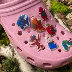 May include: A pink Croc shoe decorated with various cartoon character charms. The charms feature a blue fish, a red dinosaur, a monkey, a rabbit, a caterpillar, and a red apple car. The shoe is displayed on a bed of moss and birch bark.