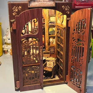 May include: A detailed miniature wooden bookshop model with open doors. The interior features tiny bookshelves, a chair, and a clock. The model has ornate cut-out designs and a sign that reads "Bookshop Memories".