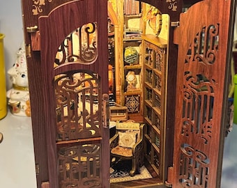 May include: A miniature wooden bookshelf model with open doors revealing a detailed interior. The model features intricate cut-out designs, a small chair, and shelves filled with tiny books. The sign above reads "Bookshop Memories".