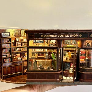 May include: Miniature Corner Coffee Shop model with detailed interior and exterior. The shop features a brick facade, wooden trim, and a sign that reads "CORNER COFFEE SHOP". Inside, there are shelves, books, and miniature furniture. The shop is lit with small lights.