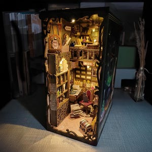 Könnte beinhalten: Ein Miniatur-Puppenhaus aus Holz mit einem detaillierten Innenraum, bestehend aus einem Bücherregal, einem Schreibtisch, einem Stuhl und einer Truhe. Das Puppenhaus ist von innen beleuchtet und schafft eine warme und einladende Atmosphäre. Das Puppenhaus ist aus Holz gefertigt und in einem dunklen Braunton lackiert. Das Puppenhaus ist mit verschiedenen Details verziert, wie z. B. einer kleinen Eulenfigur, einem Schild mit der Aufschrift "Magie" und einer kleinen Truhe.