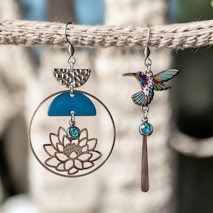 Può includere: Un paio di orecchini non abbinati. Un orecchino presenta un design geometrico blu e argento con un fiore e un cristallo blu. L'altro orecchino presenta un colibrì colorato con un cristallo blu.