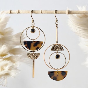 May include: Two pairs of gold-toned earrings with geometric designs. Each earring features a circular frame, a tortoiseshell-patterned half-circle, and a small black stone. One earring has a textured half-circle and a vertical bar.