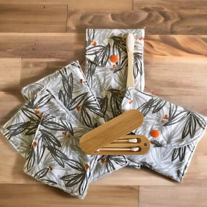 Puede incluir: Un juego de tres bastoncillos de algodón reutilizables en un estuche de bambú, junto con un cepillo de dientes de bambú y una pequeña bolsa con estampado floral. La bolsa tiene un cierre de botón y está hecha de una tela suave gris con un diseño floral blanco.